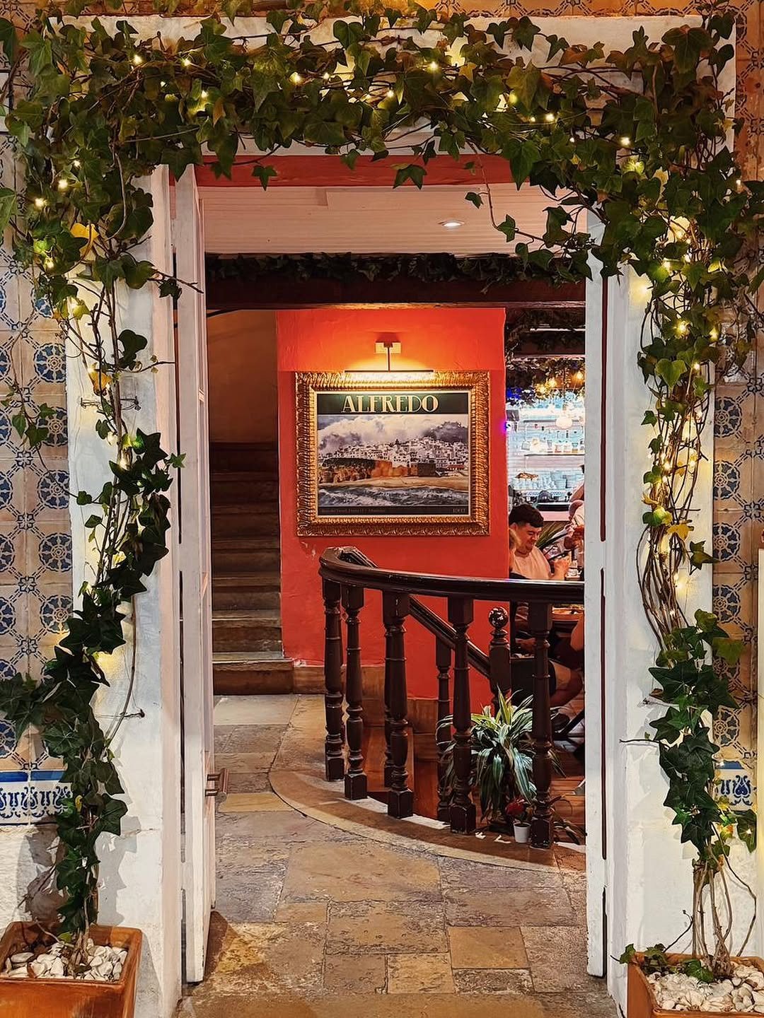 Entrada do Alfredo com um arco coberto de hera e luzes, e uma sala de paredes vermelhas ao fundo
